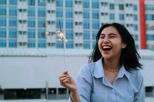 contento asiático joven negocio mujer participación bengalas a celebrar nuevo año víspera en pie en al aire libre techo parte superior con ciudad edificio antecedentes foto