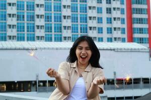 carefree asian young woman holding sparkler celebrate new year eve with laugh and dance in rooftop apartment outdoor with city building background photo