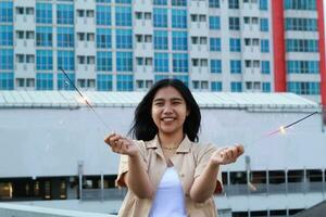 carefree asian young woman holding sparkler celebrate new year eve with laugh and dance in rooftop apartment outdoor with city building background photo