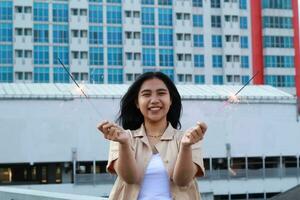 carefree asian young woman holding sparkler celebrate new year eve with laugh and dance in rooftop apartment outdoor with city building background photo