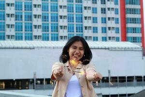 carefree asian young woman holding sparkler celebrate new year eve with laugh and dance in rooftop apartment outdoor with city building background photo