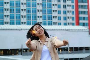 carefree asian young woman holding sparkler celebrate new year eve with laugh and dance in rooftop apartment outdoor with city building background photo
