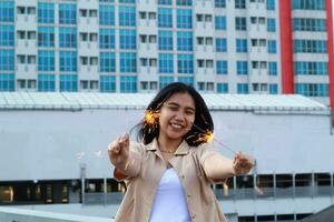 carefree asian young woman holding sparkler celebrate new year eve with laugh and dance in rooftop apartment outdoor with city building background photo