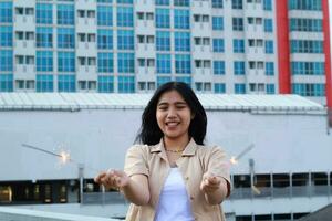 carefree asian young woman holding sparkler celebrate new year eve with laugh and dance in rooftop apartment outdoor with city building background photo