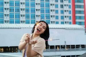 carefree asian young woman holding sparkler celebrate new year eve with laugh and dance in rooftop apartment outdoor with city building background photo