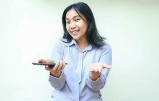 satisfied asian young business woman holding smartphone do presenting gesture with open palm to camera with smiling happy face, offering imaginary app product, wear formal shirt isolated in white photo
