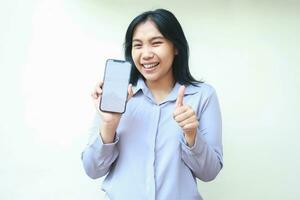 smiling happy asian young business woman giving thumbs up to imaginary product on mobile phone screen, excited female wear formal shirt laughing, isolated on white background photo