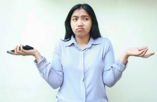 confused asian young woman holding smartphone and spreading palm with do not know gesture and have no idea, female in formal suit looking away isolated in white background photo