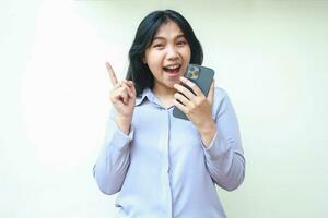 playful asian young business woman holding cell phone pointing up with excitement and smiling broadly to camera wear formal suit isolated on white background photo