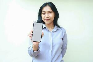 confident asian young business woman show white screen of smartphone to camera with presenting gesture, wear formal shirt standing isolated white background, product app mockup, advertisement concept photo