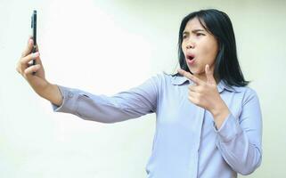 shocked asian young business woman pointing finger with thumbs up to mobile phone, stunned with wow expression, wearing formal shirt standing over isolated white background photo