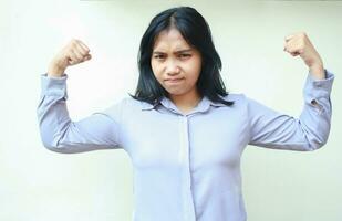 strong asian business woman raising arms showing bicep muscle strength with serious face expression looking at camera wearing formal shirt standing over white background photo