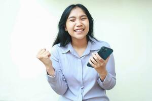 excited asian young business woman holding mobile phone and raising fist pump say yes gesture winning achievement, celebrate victory with dancing, wearing formal shirt standing over white background photo