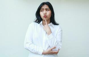 portrait of unhappy asian young business woman thinking trouble solution dressed in white formal with fingers on chin and arms folded, pensive and dreamy face expression photo