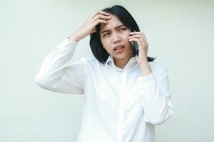 portrait of confused asian woman manager speaking on smartphone touching her forehead looking away, show regret and puzzled face expression for mistakes, wear white formal suit, standing photo