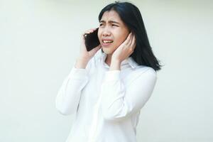 studio shot of overworked asian business woman wearing white formal shirt speaking on mobile phone covering ears from noise looking away with clueless puzzled facial expression, standing photo