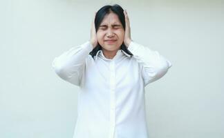 portrait of depressed asian young business woman entrepreneur covering ears with palms and closed eyes wearing white formal suit, stressed female employee under pressure, standing photo