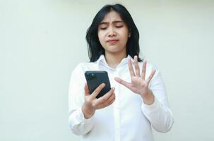 studio shot of unsure asian woman executive holding smartphone raising palms show wait gesture and afraid scared facial expression, consider offering, read news, wearing white formal shirt, standing photo