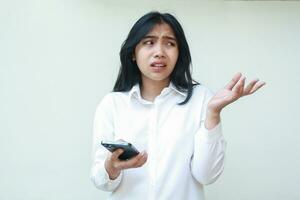 portrait of thoughtful asian business woman manager looking aside to empty space confused, raising palms showing presenting gesture, holding smartphone wearing white formal shirt, standing photo