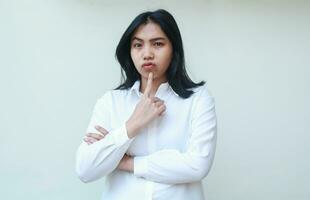 image of pensive pretty asian woman manager wearing white shirt looking at camera pondering with fingers on chin and folded arm, standing unsure expression photo