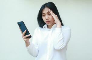 exhausted asian woman manager using smartphone holding her head suffering migraine pain, read bad news, messages, social media, overtime meeting, wear white formal suit with sleepy eyes, standing photo