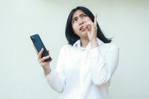 portrait of thoughtful asian office woman dark haired holding smartphone confused looking and pointing up wearing white formal shirt standing over isolated background photo