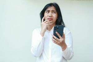 dark hair pondering asian young business woman wear white formal suit using mobile phone app looking away show worry face expression, read bad news and message, standing over isolated background photo