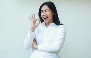 playful success asian young business woman flirting blink eye to camera giving ok signs with folded arms wearing white formal suit shirt, smiling feel satisfaction, standing over isolated background photo