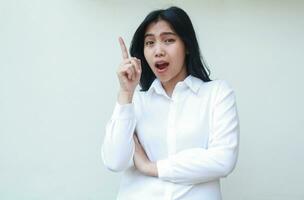 carefree asian young business woman pointing up to empty space with arms folded looking camera surprised show wow expression wearing white formal suit shirt isolated on white background photo