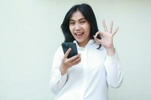 delighted playful asian young woman secretary wearing white formal suit shirt showing ok sign, okay symbol, looking camera satisfied of online shopping application, recommend gesture photo