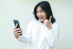 portrait of excited young asian woman entrepreneur giving thumbs up smiling playful satisfied looking at smartphone holds on hands wearing white suit shirt standing isolated, good service gesture photo