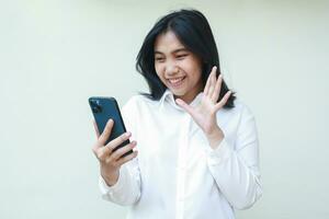 image of successful asian young business woman manager smiling excited looking at smartphone hold on hands with raising palms say hi hello gesture wearing white shirt, standing isolated photo