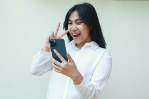 confident young asian woman holding smartphone excited taking selfie of carefree smile face expression with two fingers peace gesture wearing white suit shirt, standing isolated, wireless lifestyle photo