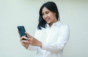 pacífico éxito joven asiático mujer vistiendo blanco traje camisa sonriente disfrutar utilizando teléfono inteligente para social medios de comunicación, en línea compras, inalámbrico comunicación chateando vídeo vocación, en pie aislado foto