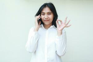 pleased satisfied asian business woman showing ok signs while speaking on smatphone wearing white formal suit shirt, approval good gesture, okay symbols, standing isolated looking at camera photo