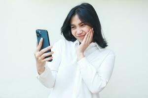grateful shy asian business woman feeling embarrassed taking selfie and video call on smartphone hold on hands wearing white formal suit shirt standing isolated, online lifestyle concept photo