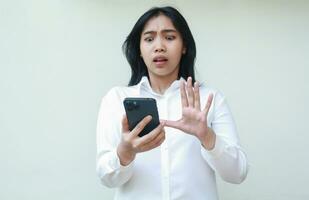 portrait of shocked asian business woman frightened looking at smartphone with raising palms showing stop gesture wearing white suit formal shirt, standing isolated, technology lifestyle concept photo