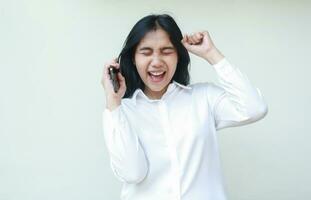 ecstatic rejoicing asian business woman celebrating winner victory by raising fist showing yes gesture while holding smartphone wearing white shirt formal suit isolated on white background photo