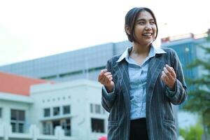 smiling asian business woman standing in city streets raising palms celebrate victory, yes gesture, wearing formal suit jacket, female executive standing in outdoors with urban building background photo