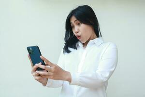 portrait of surprised asian business woman holding smartphone showing wow face expression wearing white suit formal shirt, browsing social media, texting message, using new apps, standing isolated photo