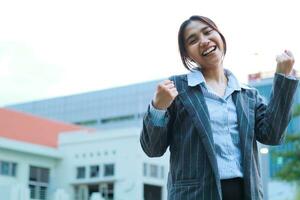 smiling asian business woman standing in city streets raising palms celebrate victory, yes gesture, wearing formal suit jacket, female executive standing in outdoors with urban building background photo