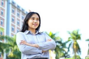 success asian businesswoman wear formal suit with cross hand standing over urban building with green plant background, ceo manager smiling confident looking away show optimistic expression photo