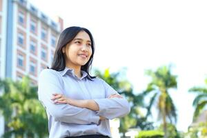 éxito asiático mujer de negocios vestir formal traje con cruzar mano en pie terminado urbano edificio con verde planta fondo, CEO gerente sonriente confidente mirando lejos espectáculo optimista expresión foto