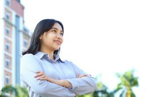 close up image of successful asian business woman standing over urban building with crossed hand wearing formal shirt, female entrepreneur smiling confident looking away show optimistic expression photo