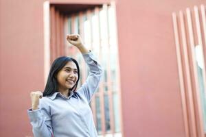 close up of smiling asian businesswoman wear formal suit celebrating winning victory standing over urban red building, female entrepreneur raising fist say yes gesture standing outdoors photo