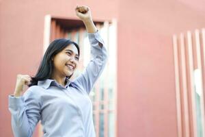 close up of smiling asian businesswoman wear formal suit celebrating winning victory standing over urban building, female entrepreneur raising fist say yes gesture standing outdoors photo