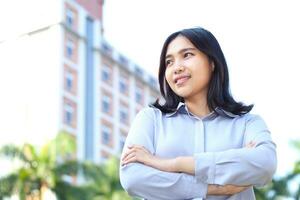 close up image of successful asian business woman standing over urban building with crossed hand wearing formal shirt, female entrepreneur smiling confident looking away show optimistic expression photo
