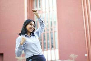 portrait of excited asian business woman standing over city building celebrate win victory wearing formal suit, indonesian saleswoman raising fist say yes gesture in outside office photo