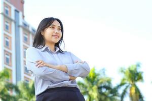 close up image of successful asian business woman standing over urban building with crossed hand wearing formal shirt, female entrepreneur smiling confident looking away show optimistic expression photo