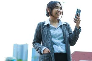 excited asian business woman holding smartphone to video call online meeting while walking on city streets with urban building background wearing formal suit photo